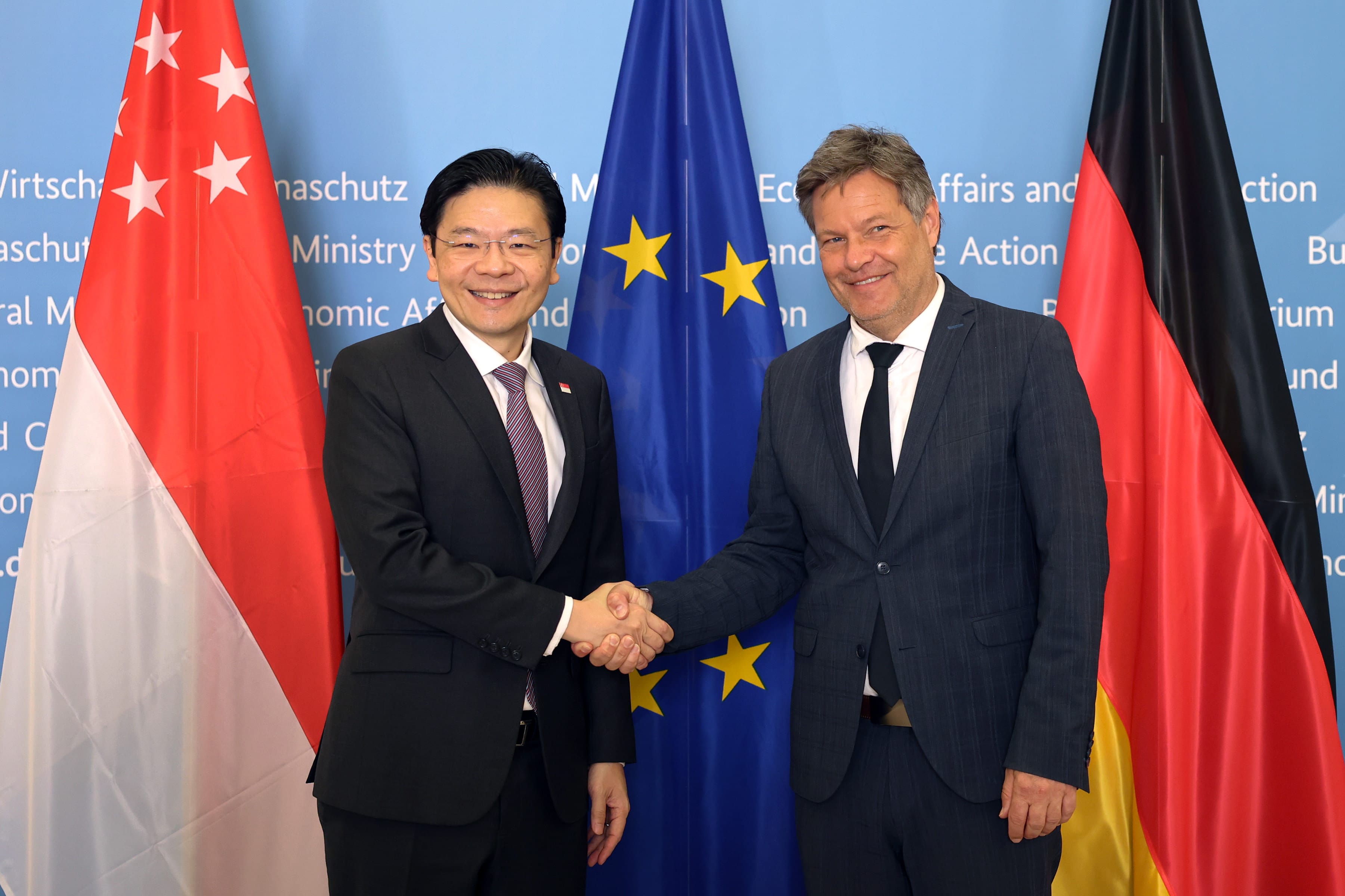 Two men in suits shake hands before the Singapore, EU, and German flags.